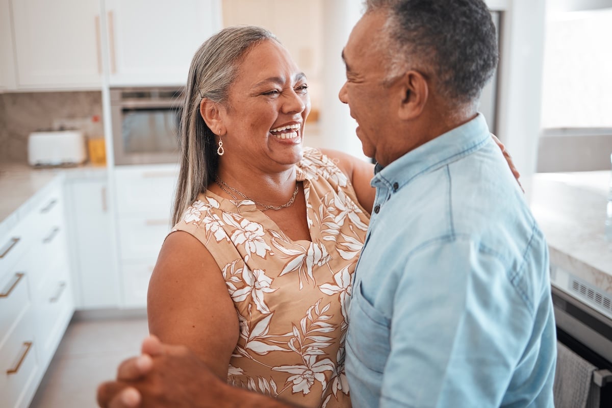 An older couple living independently at home dances in their kitchen, illustrating the freedom of aging in place.